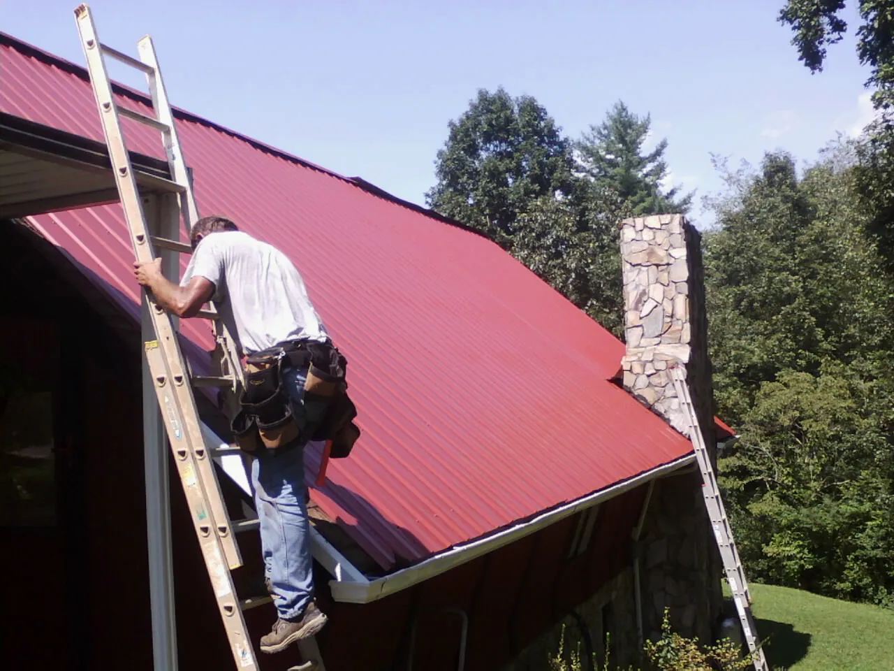 Roofer on a ladder beside a red metal roof and stone chimney — Soffit & Fascia Repair by Roofing Brothers in Asheboro
